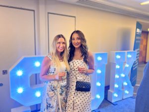 Clinical Lead for Desktop Case Management, Jane Rees, in a white and blue flowery dress with long blonde hair, stood next to Virtual Case Manager of the Year winner at the CMSUK Awards, Lydia Moore, in a white and black spotty dress, black Chanel handbag and long brown wavy hair.