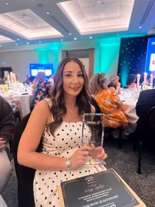 Virtual Case Manager of the Year winner at the CMSUK Awards, Lydia Moore, holding her trophy in a white and black spotty dress with long brown hair.