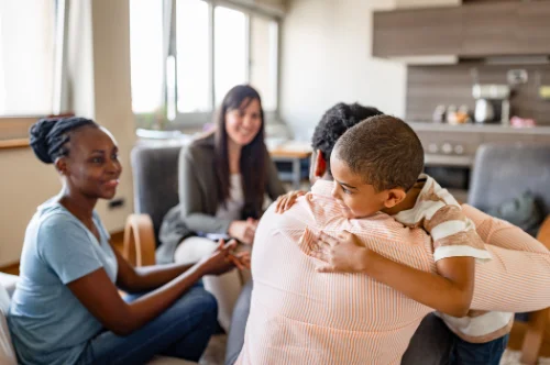A female Case Manager with long brunette hair and wearing a jacket sat in the living room of a family home opposite a Mother and Father and their young son on the soafa. The Father is embracing his son whilst the Mother and Case Manager look on fondly.
